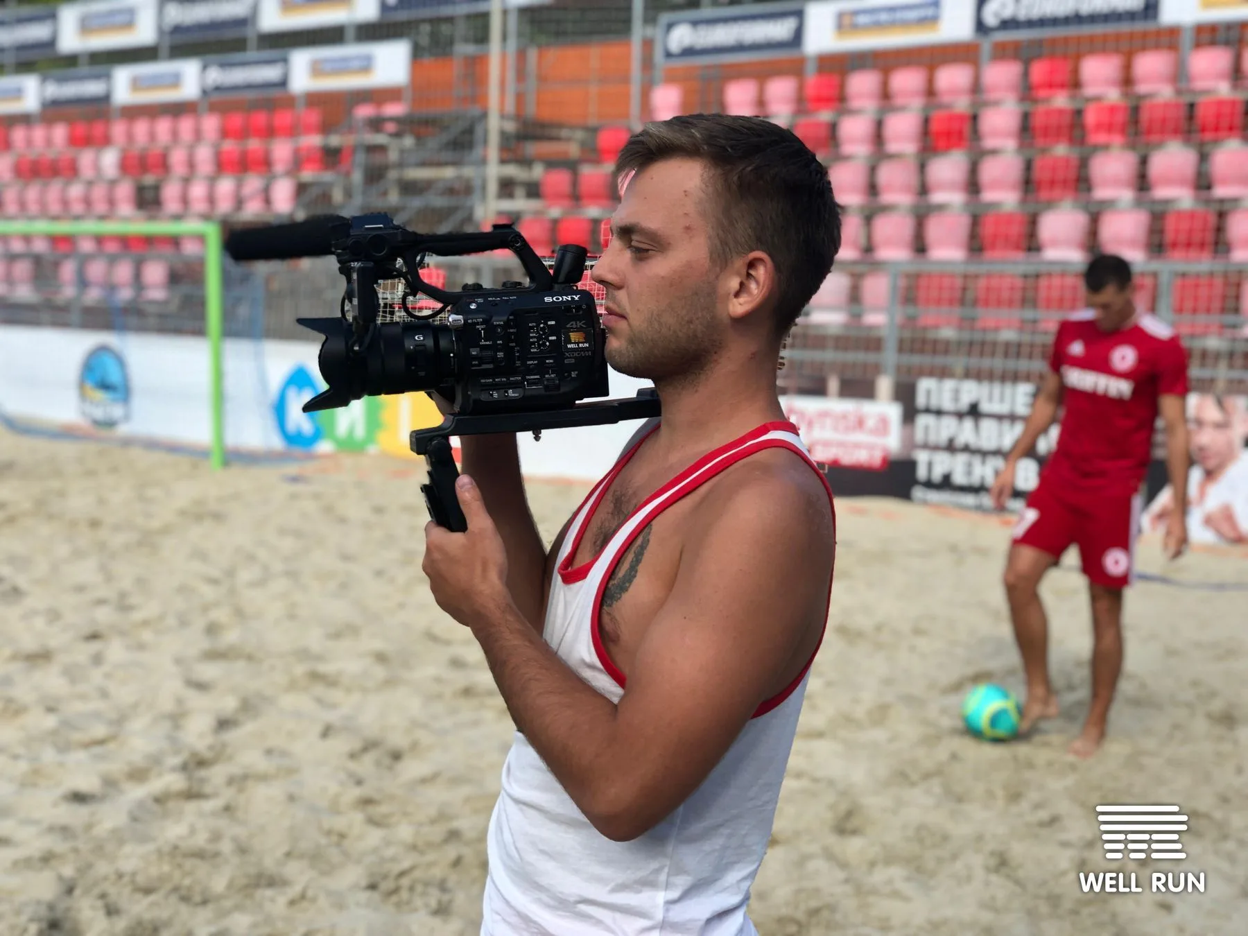 Cameraman holding Sony 4K camera on sandy beach soccer pitch, player and ball visible behind — frame 1