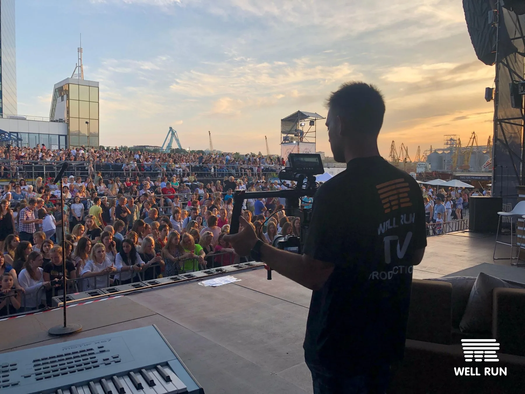 Cameraman on stage holds gimbal rig filming large outdoor concert crowd at sunset in Odessa
