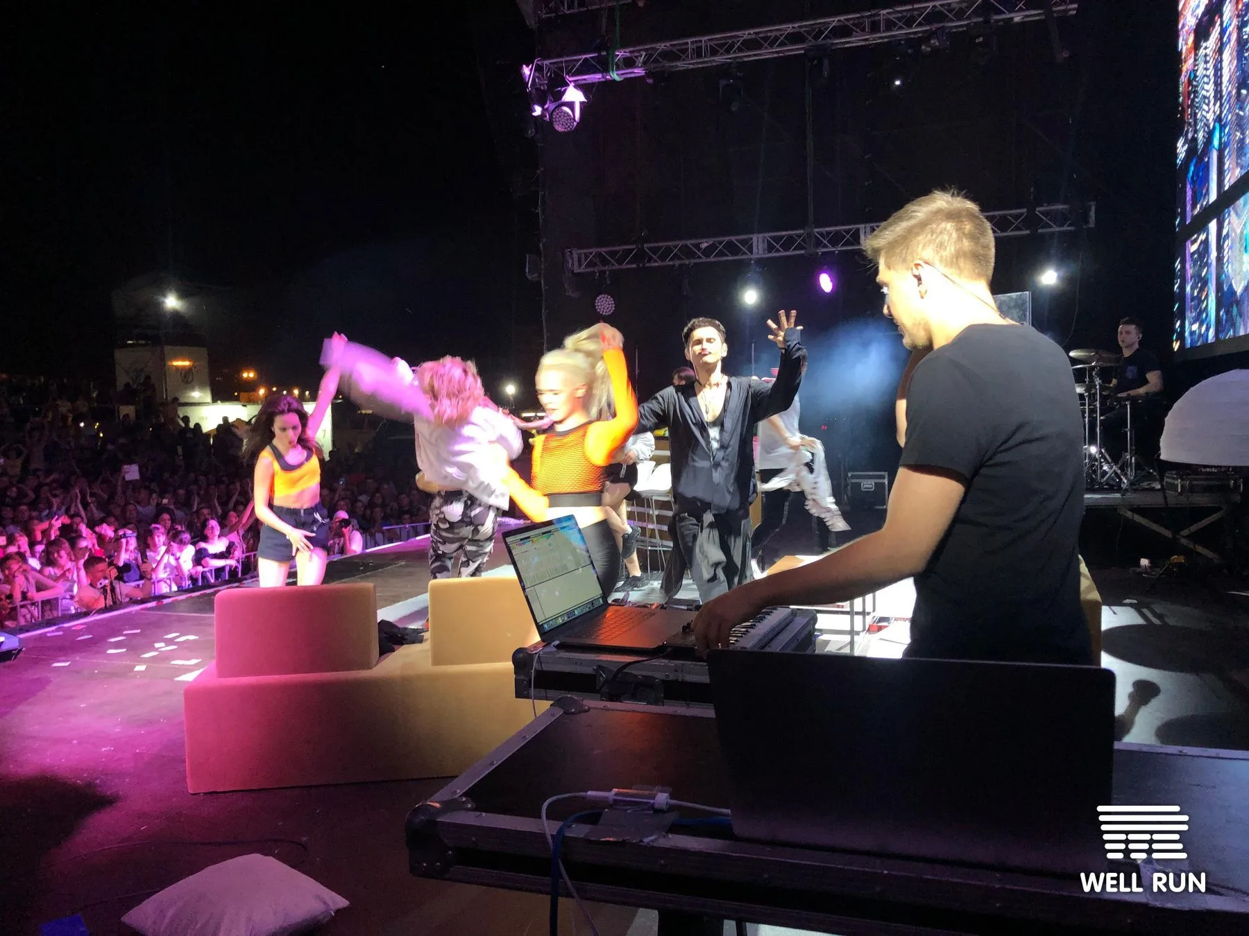 DJ operates laptop at stage-side rig as dancers and Dan Balan perform before large outdoor crowd