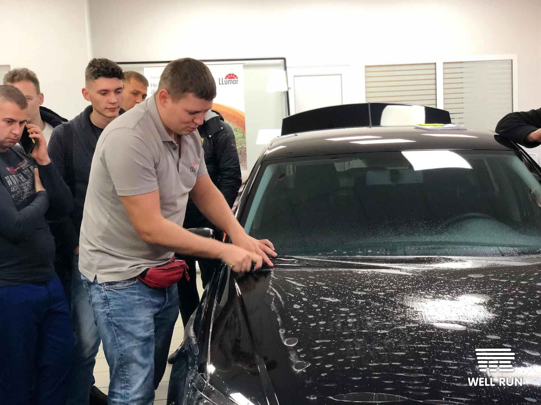 LLumar-branded instructor smoothing window tint film onto black car hood, group watching closely indoors