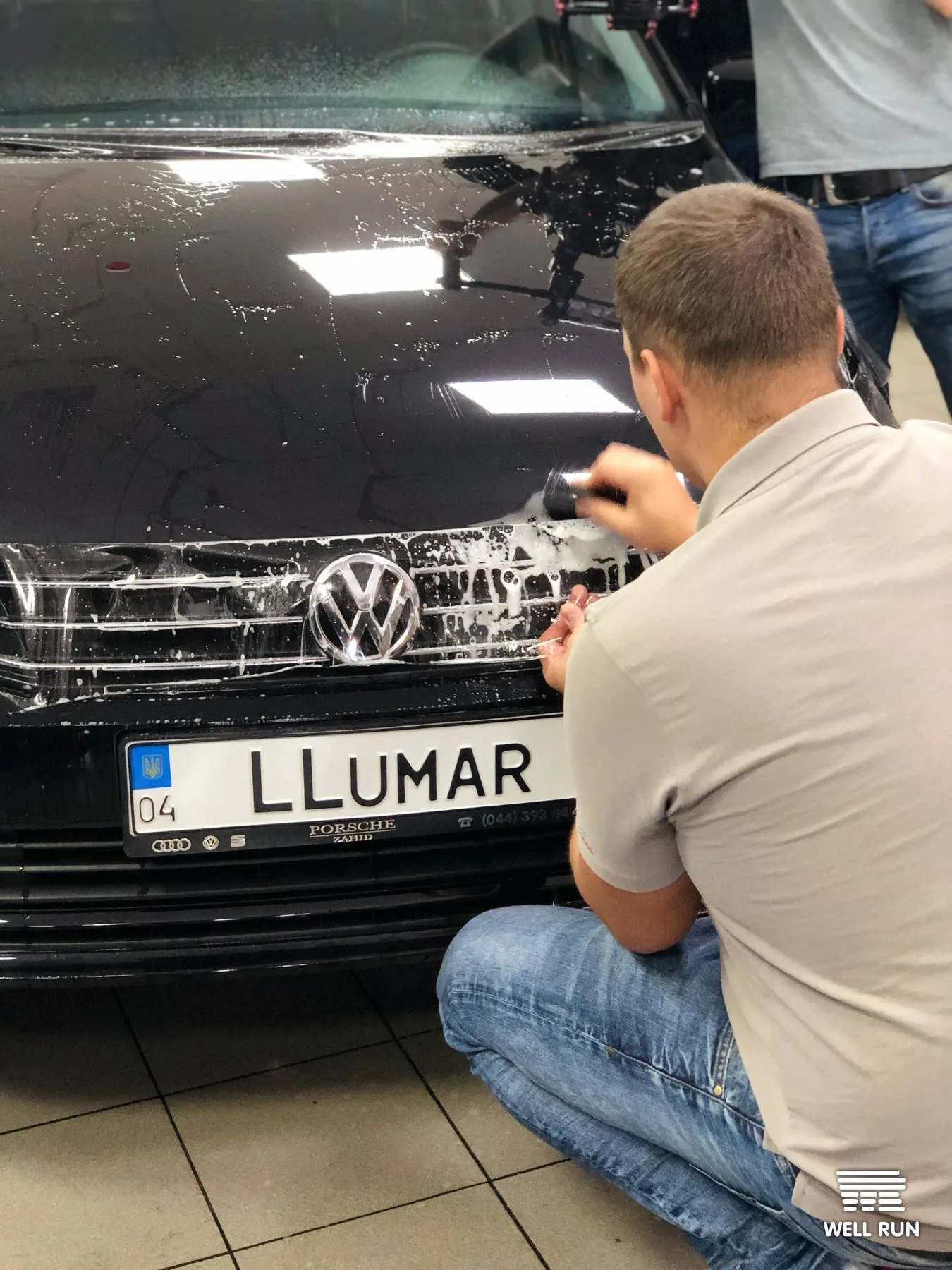 Man crouching applies soapy foam to black Volkswagen grille bearing LLumar licence plate