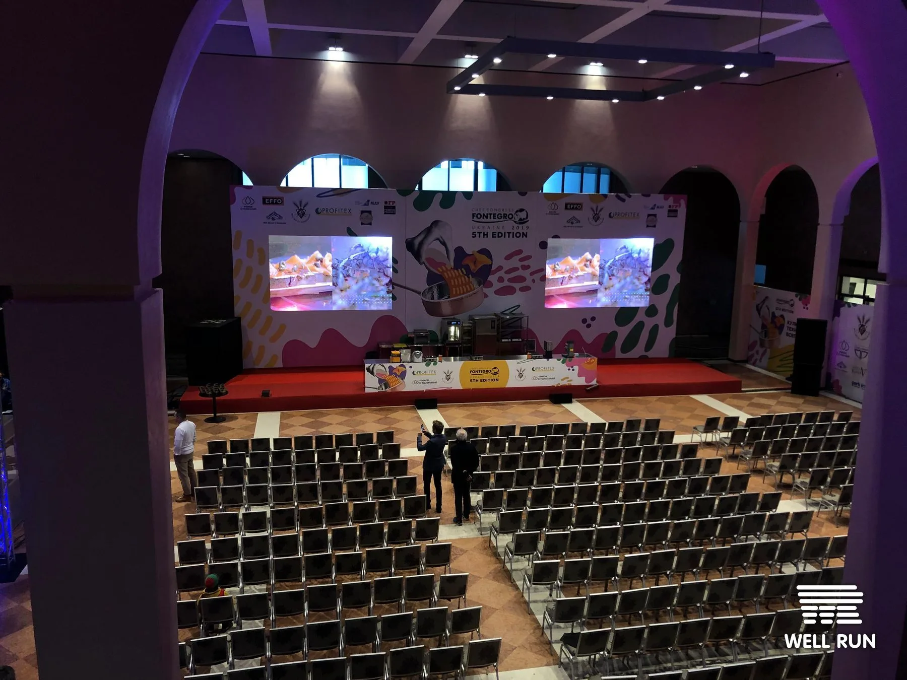 Empty conference hall with red-carpet stage, two LED screens, and rows of chairs before Fontegro Ukraine Chefs Congress