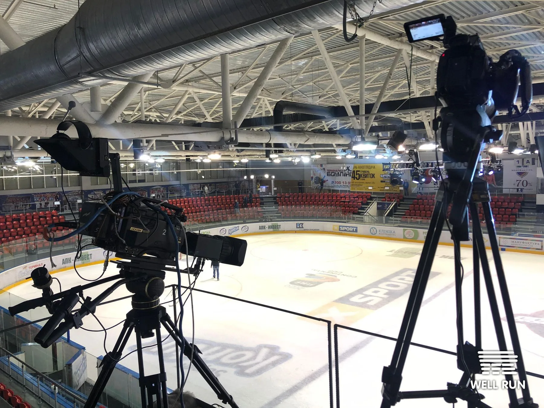 Two professional video cameras on tripods positioned high above Brovary Arena ice rink, red seats visible below — frame 1