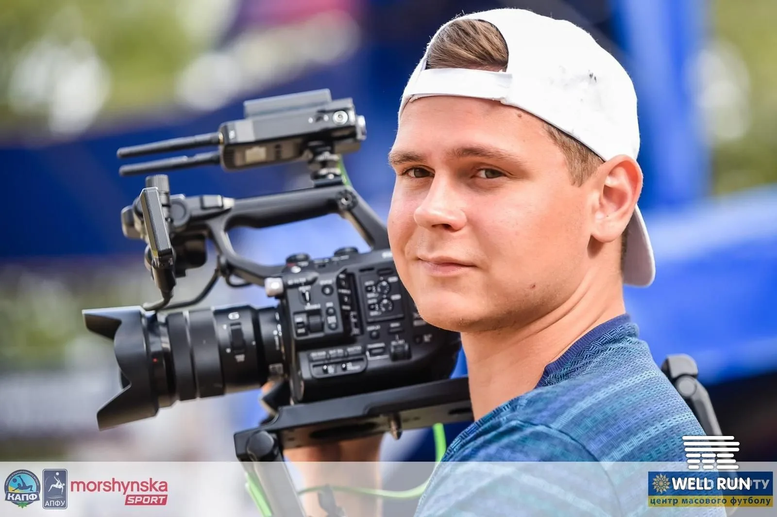 Cameraman in white cap holds Sony cinema camera rig on shoulder at outdoor sports venue