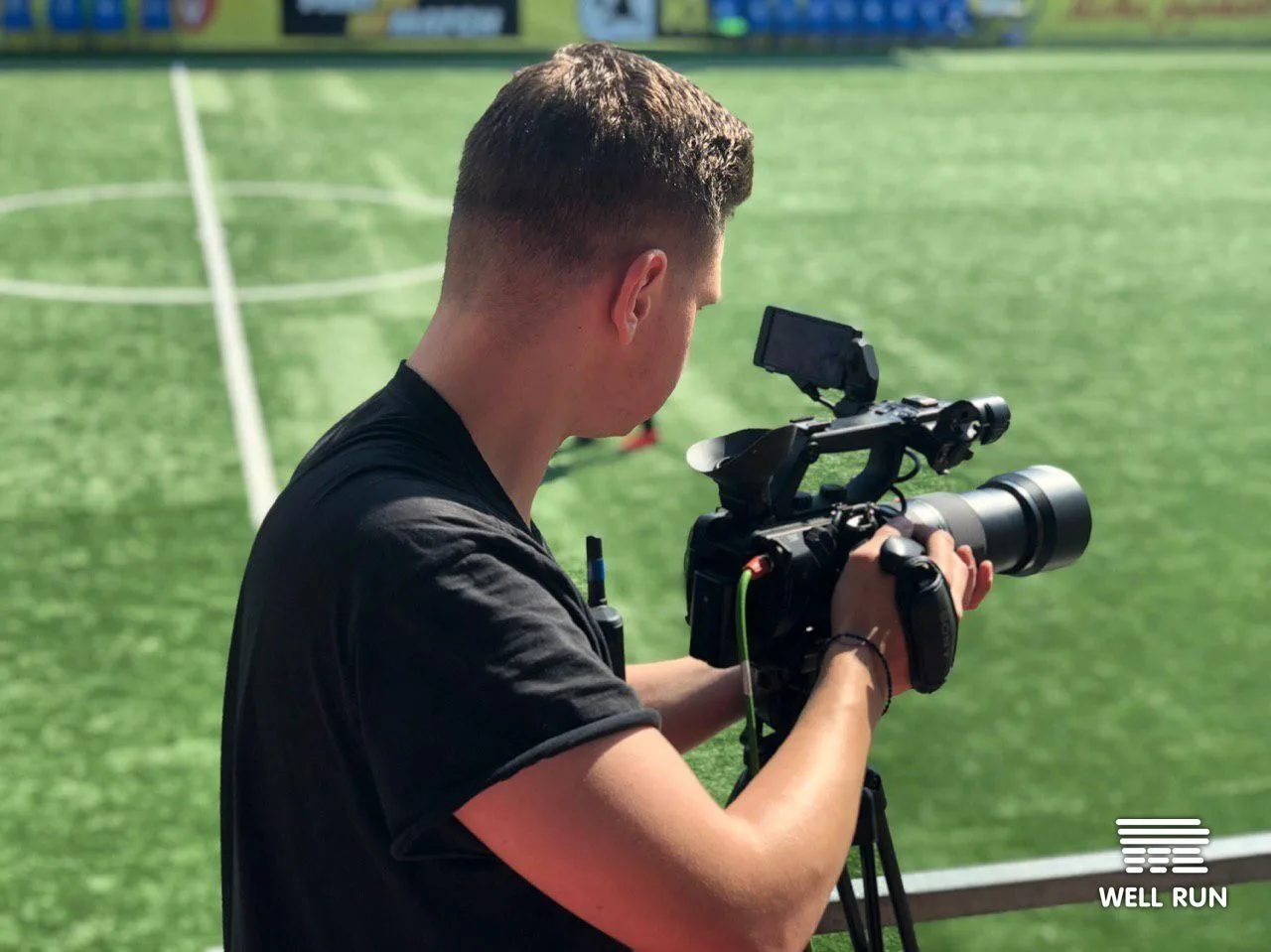 Cameraman in black t-shirt operates professional video camera on tripod at football pitch sideline
