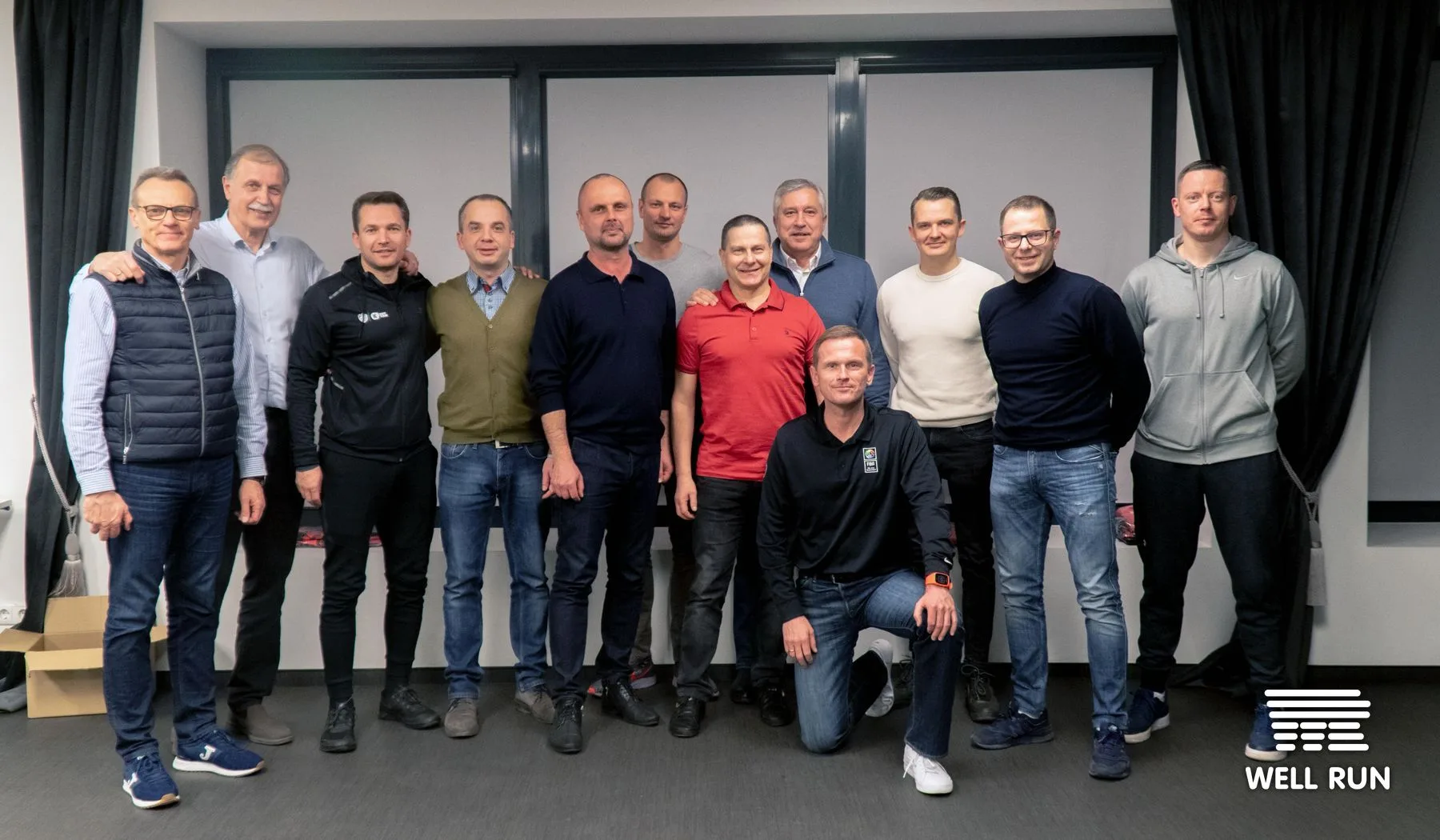 Thirteen men including Algimantas Pavilonis and Mykola Moseichenko pose together indoors at a Well Run LKL event — frame 1