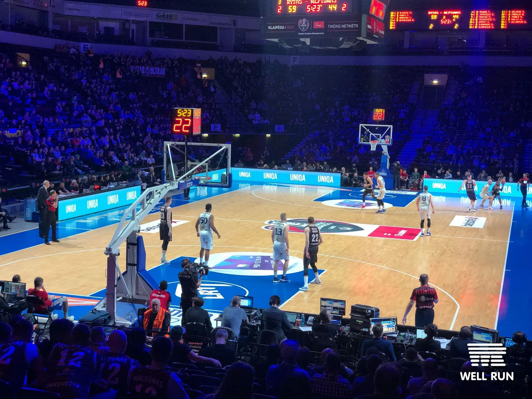 Basketball game in progress at Švyturys Arena, Klaipėda, with TV camera crew and courtside monitors visible