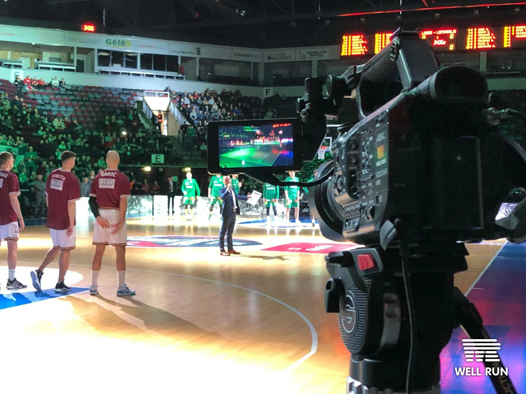 Broadcast camera on tripod captures pre-game introductions at Švyturys Arena, Klaipėda, two teams on court — frame 6
