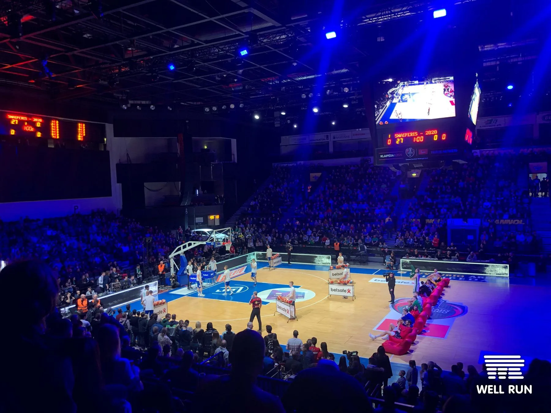 Basketball court at Švyturys Arena, Klaipėda, lit in blue with players in a three-point shootout, large scoreboard and jumbotron visible overhead, packed crowd — frame 8