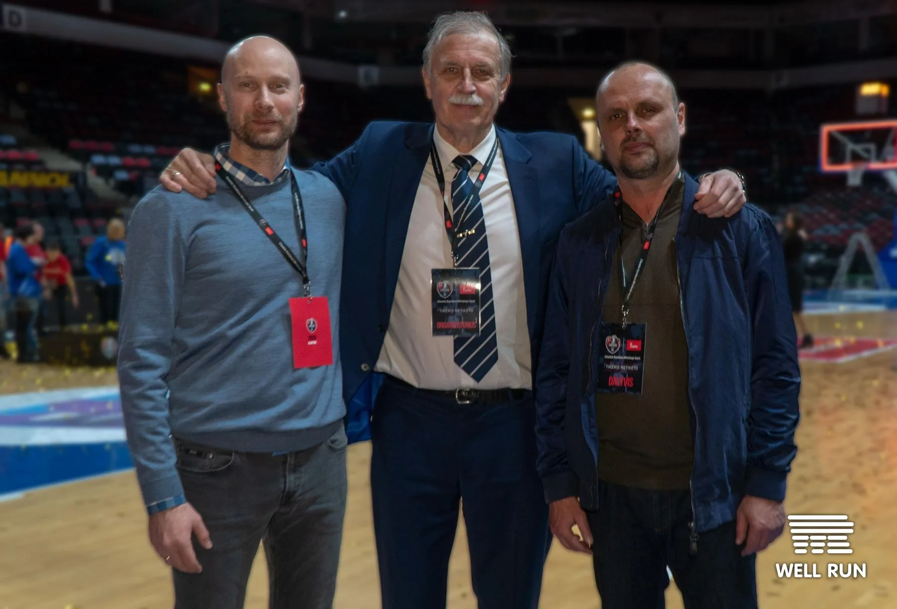 Three men with event lanyards pose on basketball court at Švyturys Arena, Klaipėda — frame 9