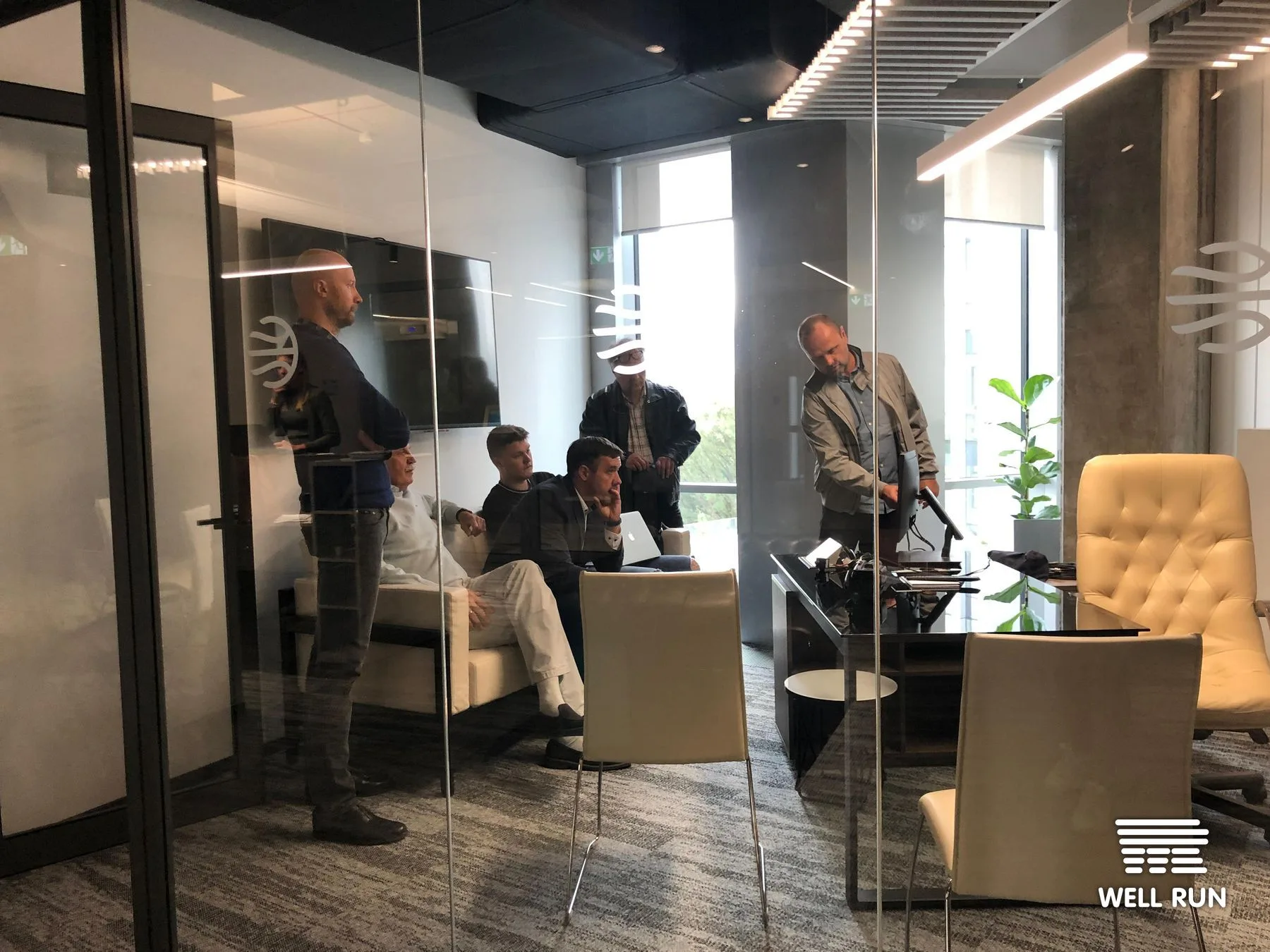 Six men in glass-walled meeting room review laptop and monitor setup at LKL headquarters Vilnius