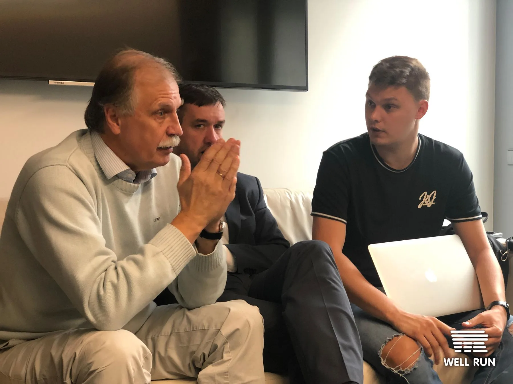 Algimantas Pavilonis gestures while two colleagues listen, laptop open, at LKL headquarters meeting