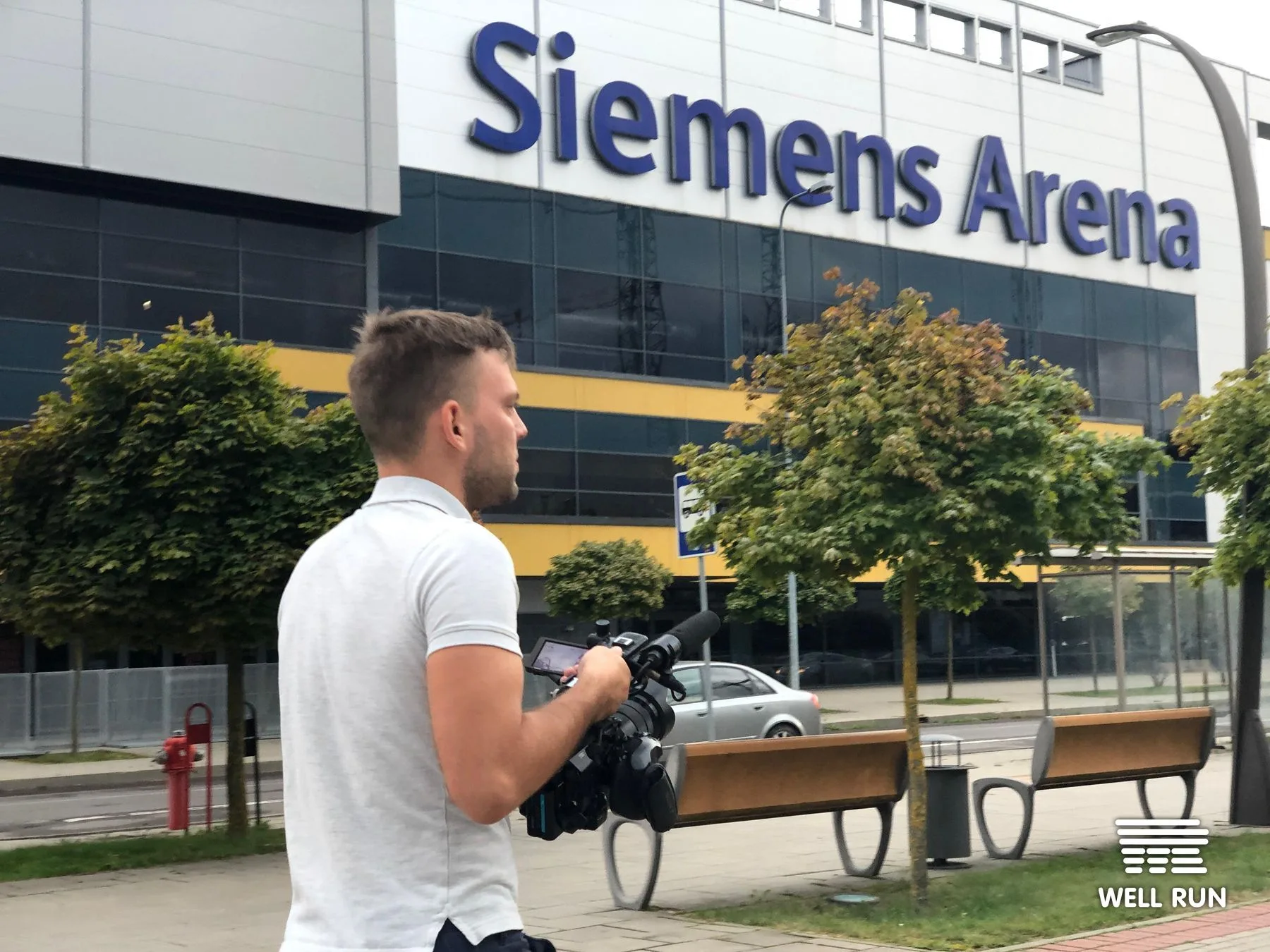 Kostiantyn Moseichenko holds video camera outside Siemens Arena, Vilnius, for LKL shoot — frame 6