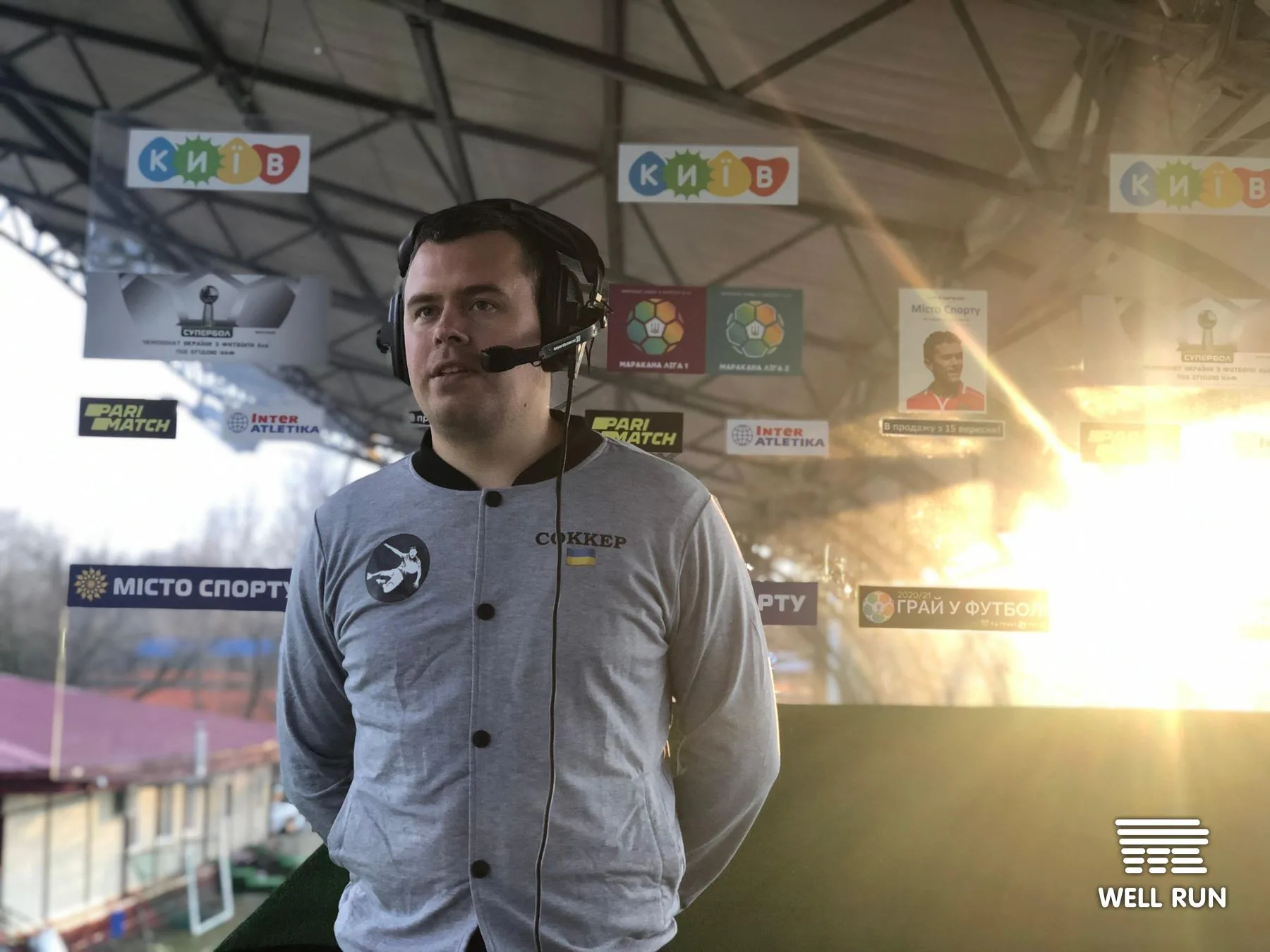 Commentator wearing headset and Socker jacket at outdoor Hydropark Arena, Kyiv, sunset behind