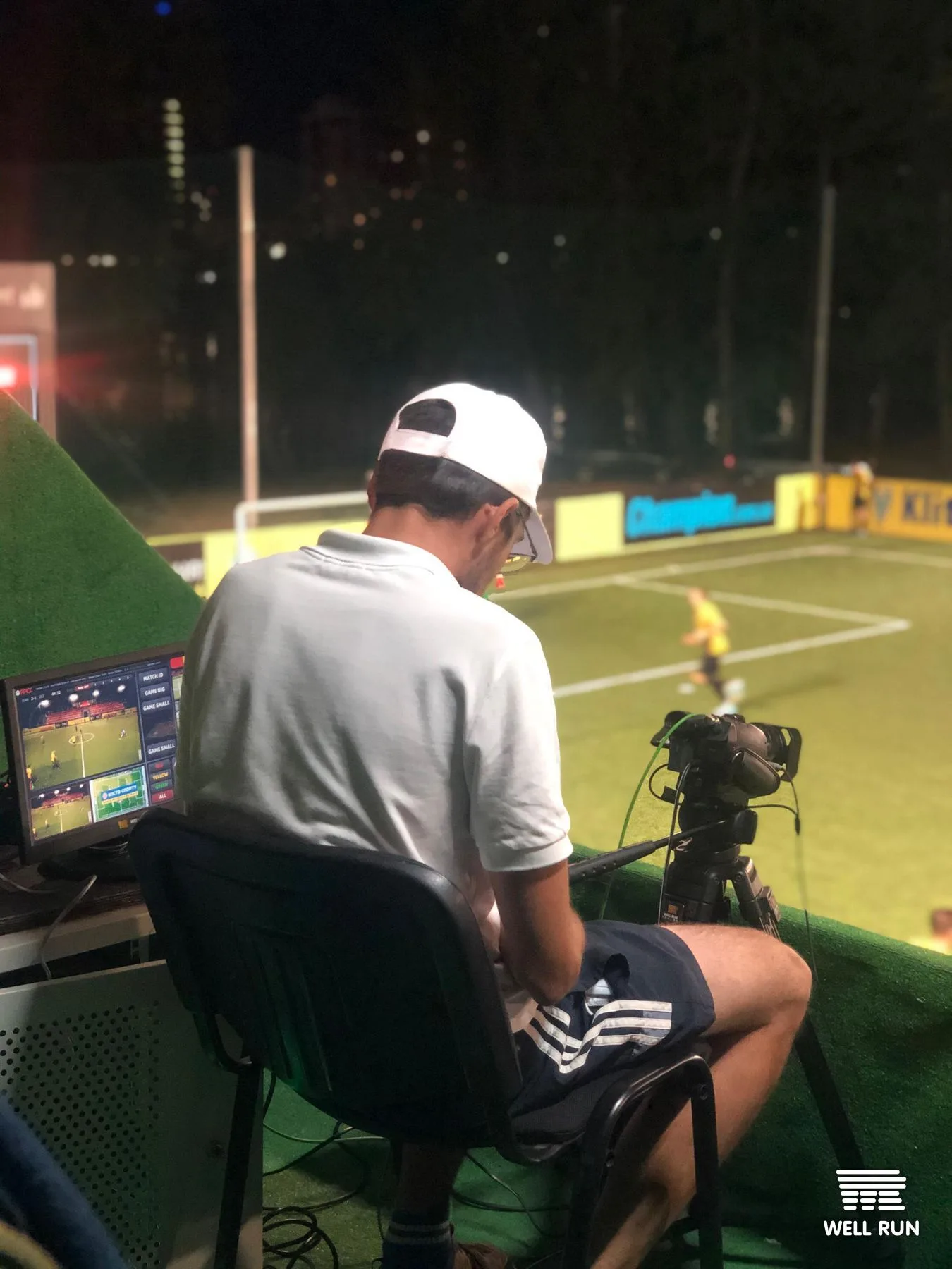 Camera operator seated at outdoor pitch-side desk, monitoring broadcast screen beside tripod-mounted camera
