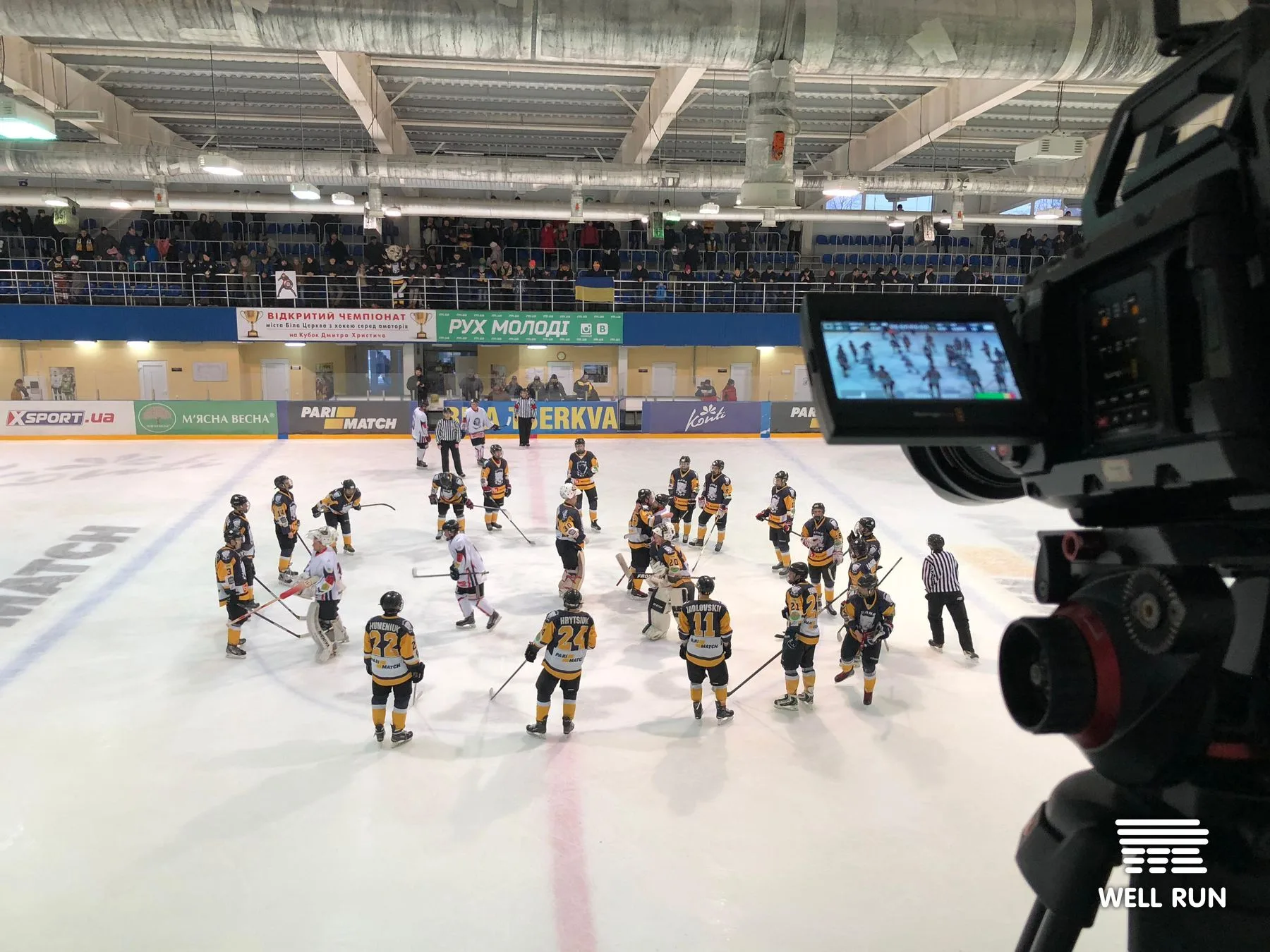 Blackmagic URSA camera on tripod filming hockey players gathered on ice at Ice Arena Bilyi Bars — frame 1